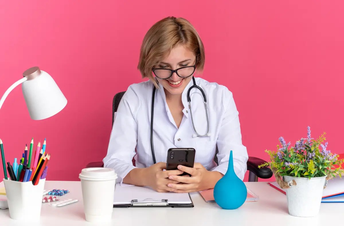 Smiling young female doctor wearing medical robe with stethoscope and glasses sits at desk with medical tools holding and looking at phone isolated on pink background